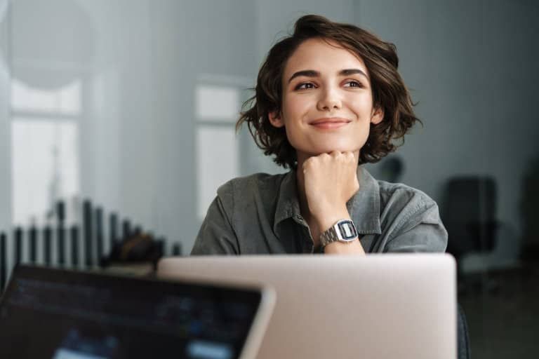 Dark-haired woman sitting behind an open laptop, smiling, with her chin propped up by her fist