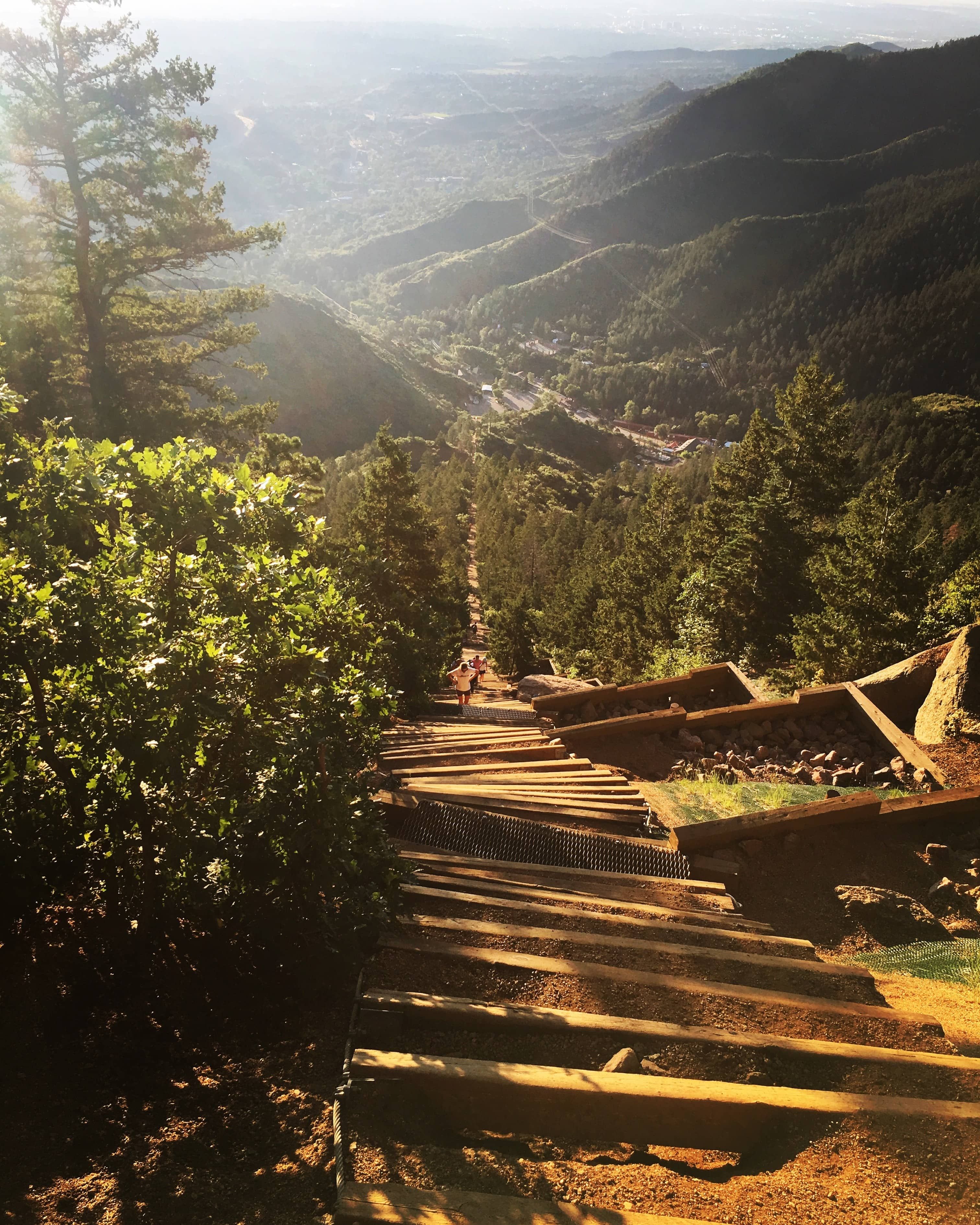 Phot of the Manitou Incline, taken about 2/3 of the way up, facing down towards the bottom. Some mountains and a little bit of Manitou Springs and Colorado Springs are visible in the background.